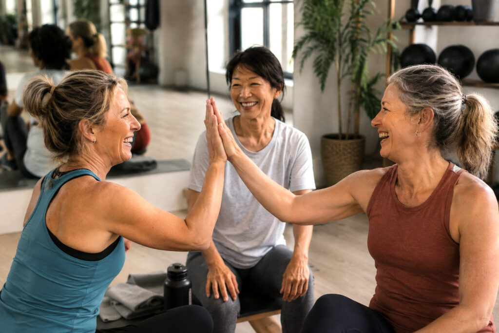 Three women in their late 40s participate in a fitness class, with two women sharing a celebratory high-five after a workout while a third smiles and looks on.