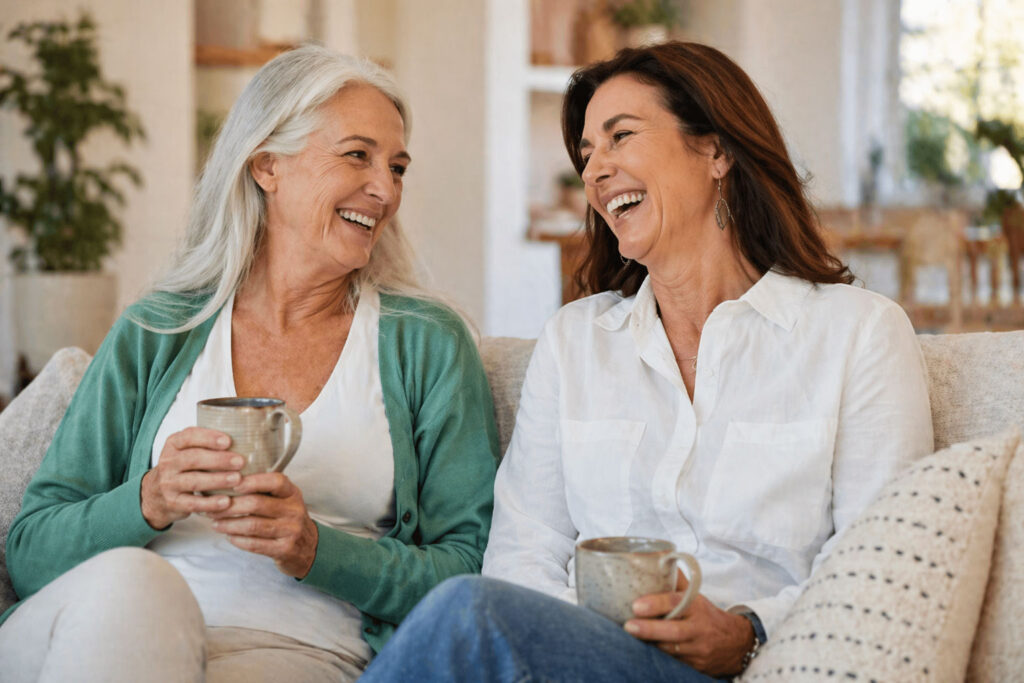 Two middle-aged women sitting together on a couch, smiling and laughing while drinking coffee in a bright, cozy home setting, reflecting warmth, connection, and everyday wellness.