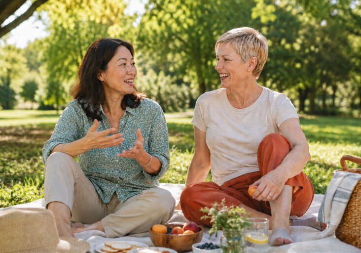 Two women in late 40s enjoying a summer picnic on a grassy lawn, with a Chinese woman gesturing as she talks and a blonde woman listening attentively surrounded by trees